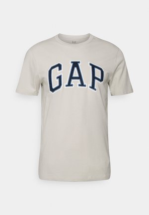 Vaaleanbeige lyhythihainen t-paita, jossa on näkyvästi esille ommeltu tummansininen "GAP"-logo rinnassa. Sujuva puuvillakankaan pinta.