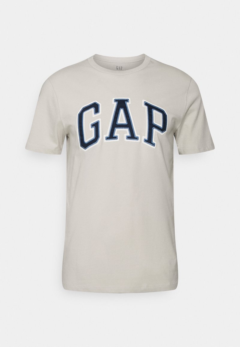 T-shirt beige à manches courtes avec un logo "GAP" bleu marine en broderie sur le devant. Texture en coton lisse.