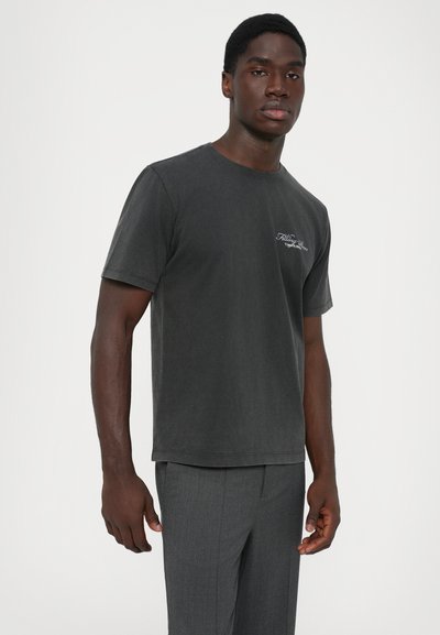 Bărbat îmbrăcat cu un tricou negru uzat, cu text alb cursiv, și pantaloni gri din stofă, stând în fața unui fundal alb simplu.