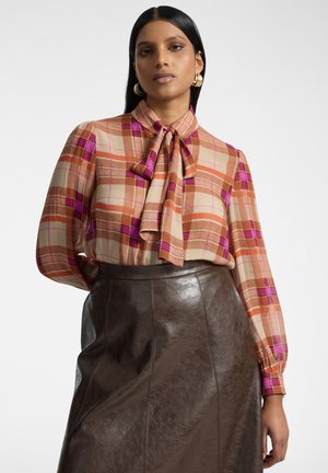 Mujer con cabello largo y oscuro, que lleva pendientes de aro dorados, una blusa de cuadros rosa y naranja con lazo en el cuello y una falda de cuero marrón oscuro.