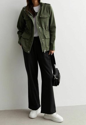 Personne portant une veste vert olive, une chemise blanche, un pantalon noir à jambes larges, des baskets blanches, tenant un sac noir sur un fond uni.