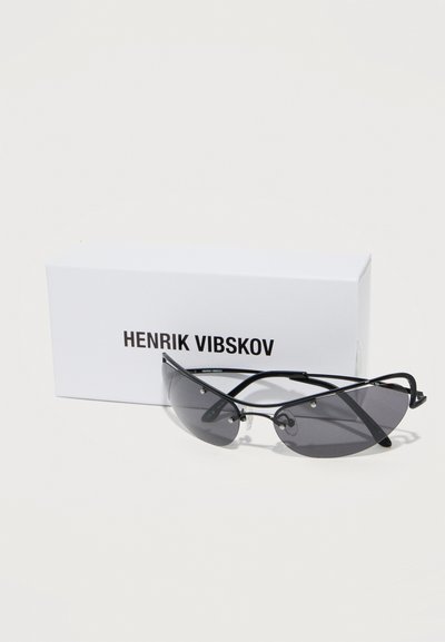 Μαύρα γυαλιά ηλίου τύπου cat-eye με σκουρόχρωμο φακό, τοποθετημένα μπροστά από ένα λευκό κουτί με την επιγραφή «HENRIK VIBSKOV» σε απλό φόντο.