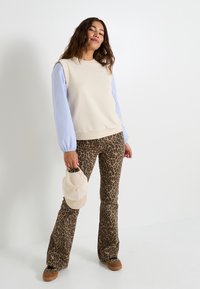 Suéter beige claro con mangas acanaladas azules, combinado con pantalones acampanados de estampado de leopardo marrón. La modelo sostiene una gorra beige.