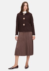 Brauner, strukturierter Cardigan mit drei blumigen Knöpfen, kombiniert mit einem gefalteten braunen Rock und glänzenden burgunderfarbenen Ballerinas. Darunter ein weißes Hemd mit Kragen.