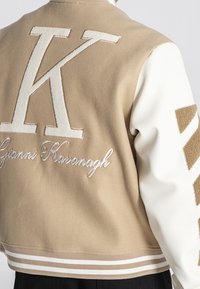 Tamnobijela bomber jakna s velikim vezenim slovom "K" i "Gianni Kavanagh", s prugastim rebrastim manžetama i dijagonalnim detaljima na rukavima.