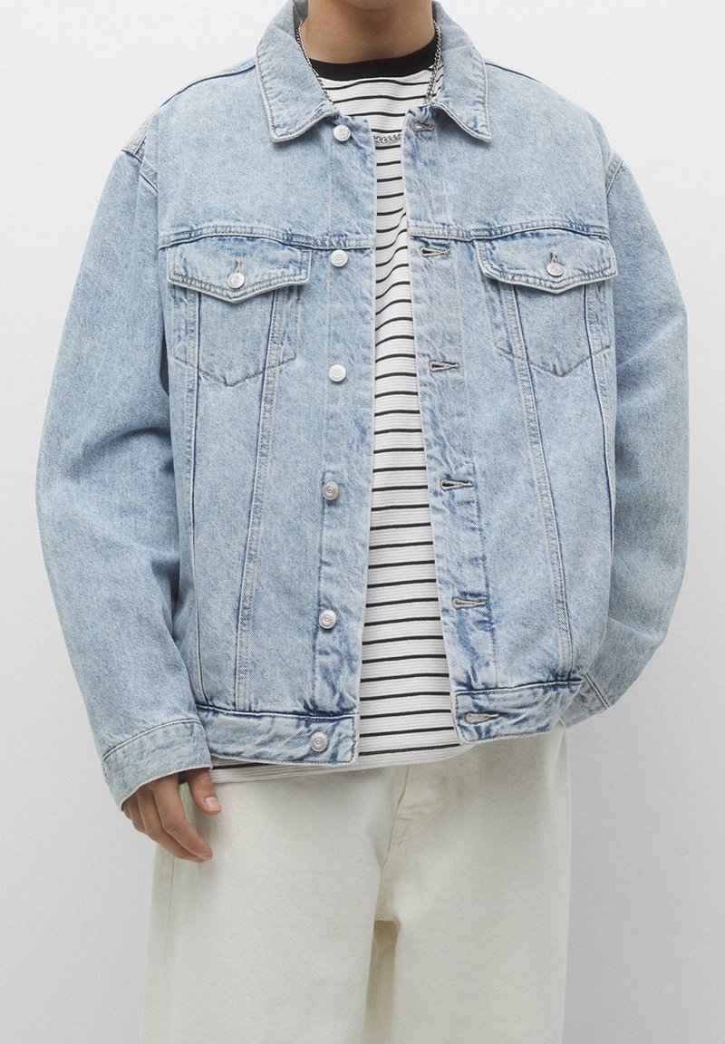 Personne portant une veste en denim bleu clair par-dessus un t-shirt à rayures noir et blanc avec un pantalon blanc cassé, debout devant un fond uni.