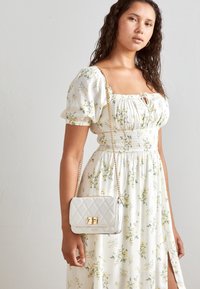 Bílé květinové maxi šaty se žlutými květy, se širokými rukávy a nařaseným živůtkem. Kvitovaná bílá kabelka s zlatými detaily.