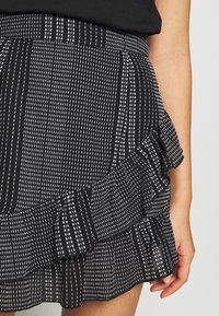 Jupe noire à volants avec un motif texturé à pois blancs. Taille élastique et design superposé, mettant l'accent sur des lignes horizontales et verticales.