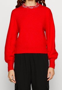 Henkilö, joka pitää yllä kirkkaan punaista villapaitaa, jossa on koristellut kaulus, ja mustia housuja, kädet rentoutuneina sivuilla.