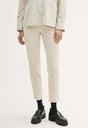 Person iført beige bukser, lyse beige langærmet skjorte med knapper, sorte sokker og sorte chunky loafers, der står på en hvid baggrund.