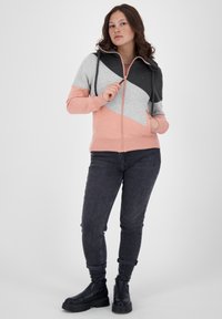 Giovane donna con capelli ricci che indossa un hoodie con zip rosa, grigio e nero, jeans scuri e stivali neri, in piedi su uno sfondo bianco.