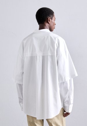 Camisa blanca de gran tamaño con mangas cortas, cuello y detalle en el yugo trasero; fabricada en algodón suave; combinada con pantalones beige.