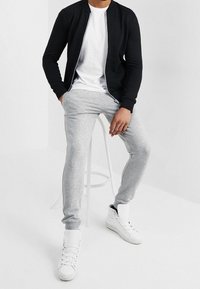 Svart zip-up bomberjacka över en vit t-shirt, kombinerad med ljusgrå sweatshirtbyxor och vita sneakers. Släta tyger och enkel design.