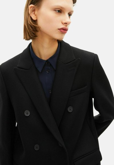 Blazer nero doppiopetto con grandi bottoni, revers a scollo e taschino sul petto. Indossato sopra una camicia colletto blu navy.