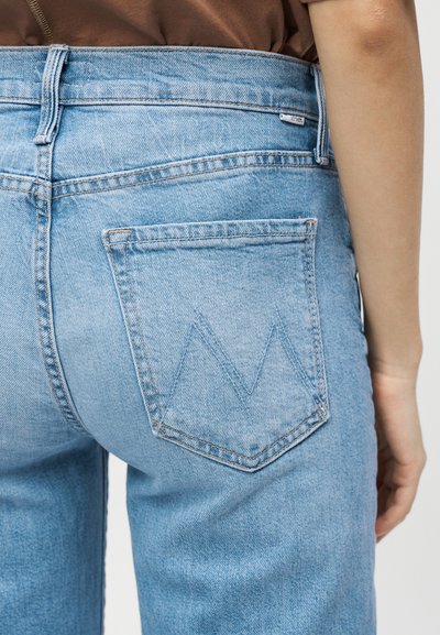 Φωτεινά μπλε τζιν με ραμμένο μοτίβο "M" στην πίσω τσέπη, φορεμένα από άτομο με καφέ μπλούζα, εμφανιζόμενα από τη μέση και κάτω.