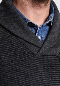 Zbliżenie na mężczyznę w ciemnoszarym swetrze z prążkowanego materiału z kołnierzem szalowym, nałożonym na zapinana niebieską koszulę dżinsową.