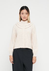 Lys rosa strikket genser med bølget krage, knappelukking og ribbestruktur. Kombinert med mørke striperbukser.