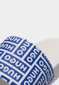 Sandale à glissière blanche avec une large lanière bleue arborant un motif répété du logo "HUGO" en lettres blanches audacieuses.