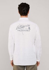 Camisa de lino blanca de manga larga con un diseño gráfico en la parte posterior que presenta "Le Soleil d'été", un icono de palmera y "FAÇONNABLE" en azul.