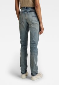 Vaqueros de mezclilla azul claro con corte recto, bolsillos traseros y detalles de costura sutiles, combinados con calzado beige sobre un fondo blanco.