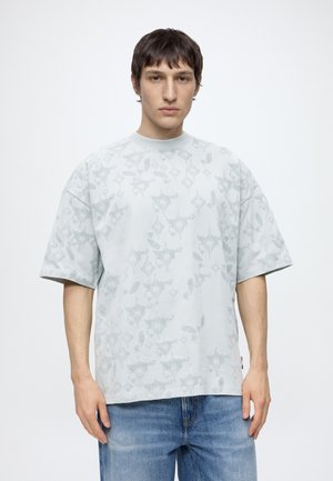 Ung mand med kort mørkt hår iført en lysegrå mønstret oversized t-shirt og blå jeans, stående mod en ensfarvet hvid baggrund.