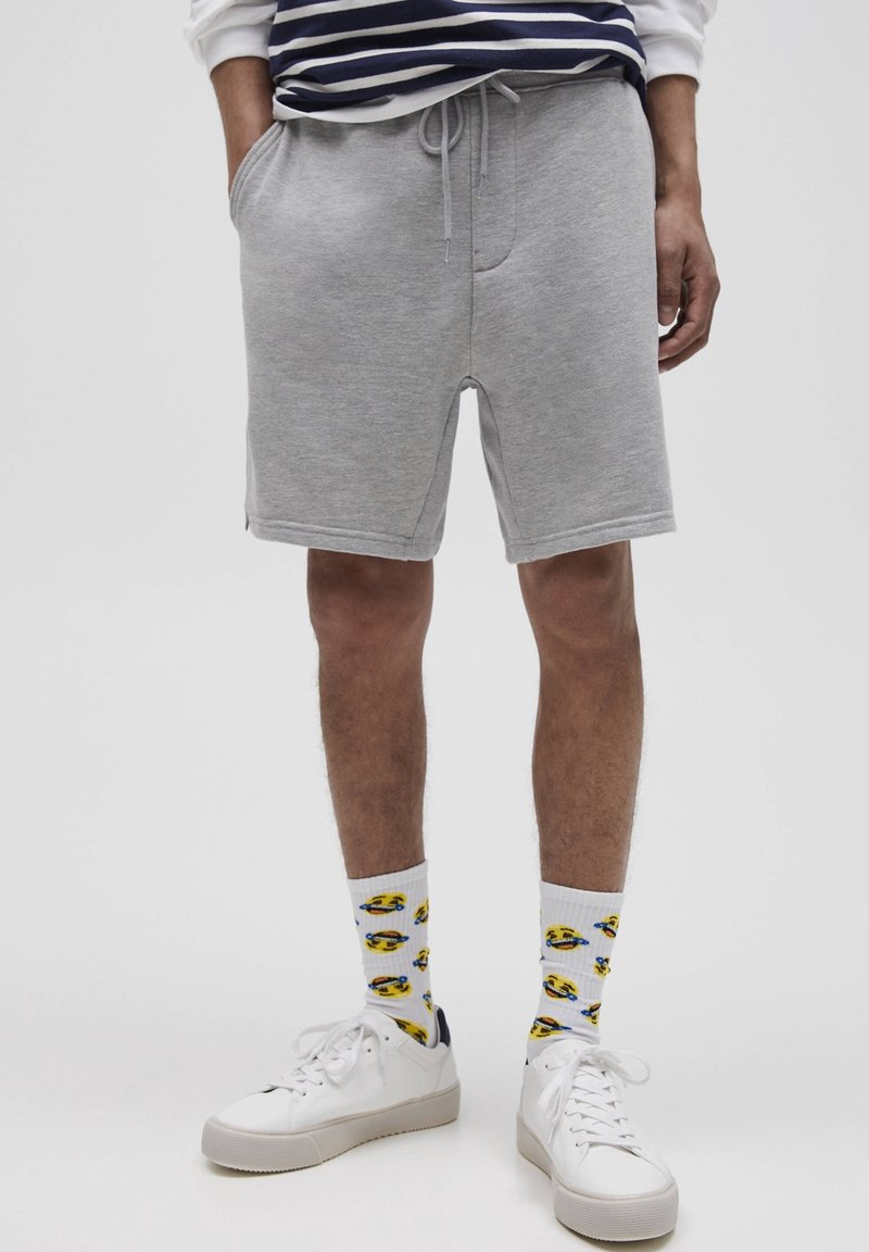 Shorts en mélange coton gris avec une taille élastique et un cordon de serrage, dotés de poches latérales. Associés à des baskets blanches et des chaussettes à motifs.