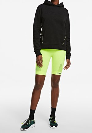 Person iført en sort hættetrøje, neongrønne cykelshorts med logo, sorte sokker og sort-grønne sportssko på hvid baggrund.