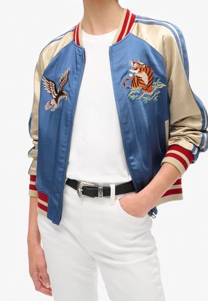 Persona con una chaqueta bomber de satén azul con águila y tigre bordados, camiseta blanca, pantalones blancos y cinturón negro con hebilla plateada.
