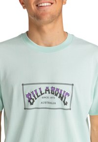 Světle modré bavlněné tričko s centrálním grafickým prvkem, na kterém je "BILLABONG" v purpurové a černé barvě, umístěné v obdélníkovém rámečku.