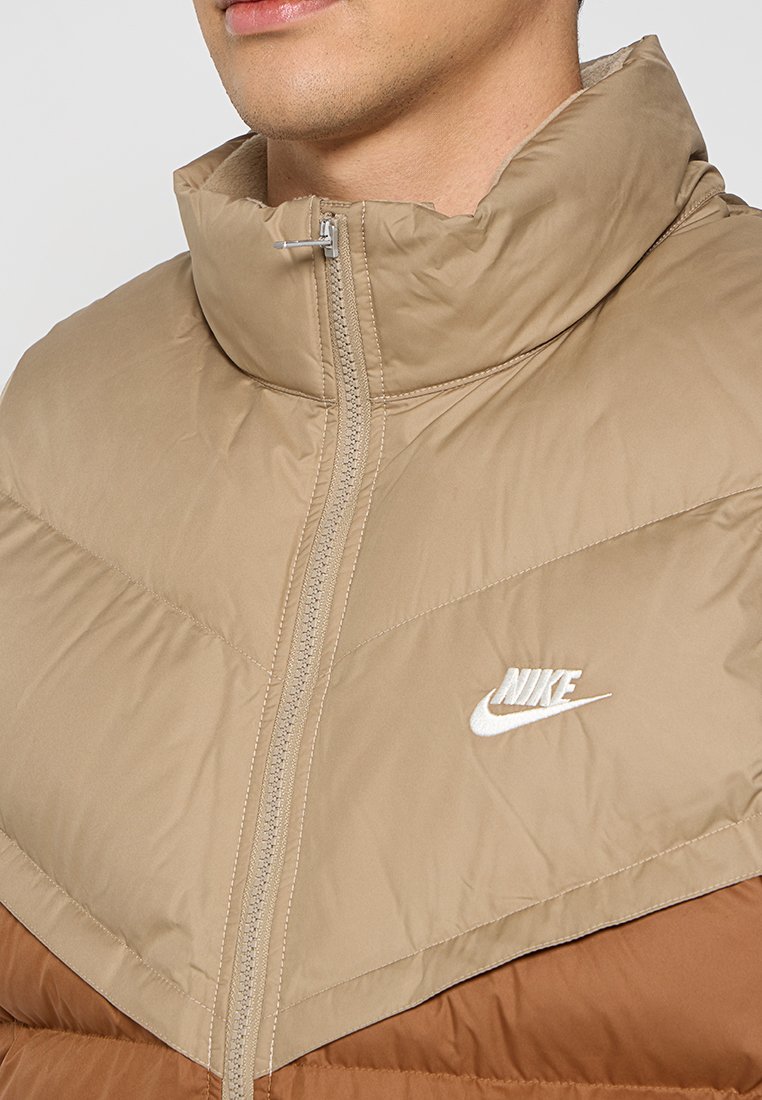 Veste rembourrée avec un col haut dans des tons beige et marron, dotée d'une fermeture éclair et d'un logo Nike blanc sur la poitrine.