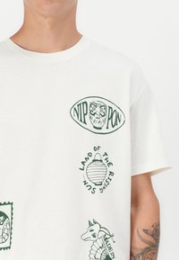 Kurzärmliges cremefarbenes T-Shirt mit grünen Druckmotiven, darunter eine Eule, eine Laterne und ein Hund, mit dem Text "Nippon" und "Land der aufgehenden Sonne."