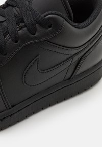 Svart sneaker i slät syntetisk läder med strukturerad gummisula. Har runda svarta snörning och en sydd Nike-logga på sidan.