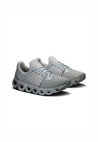 Des chaussures de sport grises avec une tige en tissu texturé, dotées d'une large languette en mesh, de lacets plats et d'une semelle extérieure rembourrée et segmentée.