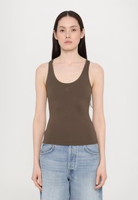Top in cotone marrone con scollatura a forma di barca e spalline larghe, realizzato in tessuto liscio. Presenta un piccolo logo al centro. Indossato con jeans azzurri chiari.