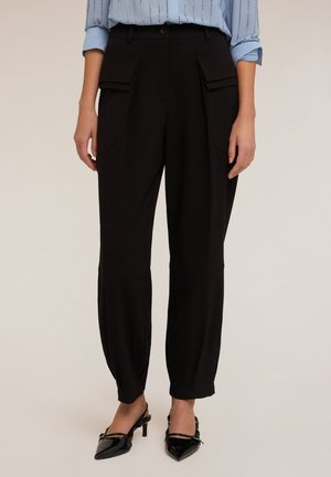 Vrouw in zwarte high-waist cargobroek met klepzakken, gecombineerd met een lichtblauw langemouwen shirt en zwarte spitse slingback hakken.