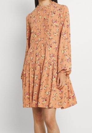 Femme portant une robe florale pêche à manches longues avec un col à volants et une jupe froncée, debout devant un fond uni.