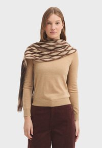 Pull marron clair avec un col rond, associé à un pantalon en velours côtelé bordeaux. Un châle à motifs en crème et marron foncé drape sur les épaules.