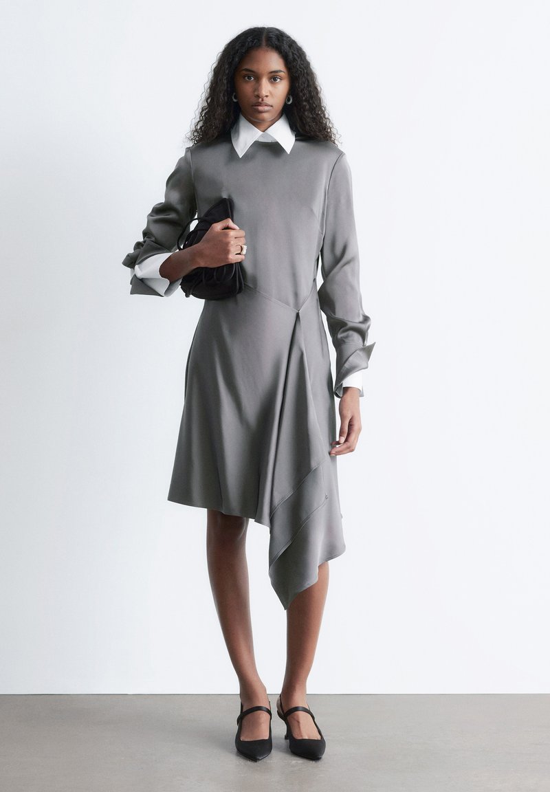 Robe grise asymétrique à texture lisse, avec un col blanc et des manches retroussées. Portée avec un sac à main noir et des chaussures noires.