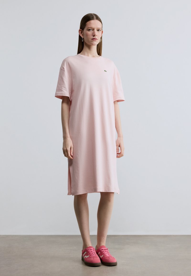 Ροζ βαμβακερό φόρεμα t-shirt με κοντά μανίκια, άνετη εφαρμογή, πλευρικές σχισμές και διακριτικό λογότυπο στο στήθος. Συνδυασμένο με ροζ αθλητικά παπούτσια.