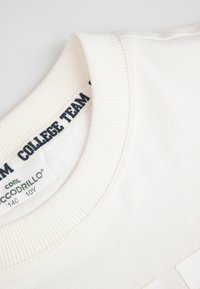 Kremowy sweter z ribbowanym kołnierzem. Posiada metkę z napisem "ZESPÓŁ UCZELNIANY". Gładka faktura, minimalistyczny design i brak wzorów.