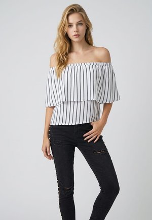 Mujer rubia con top blanco sin hombros con rayas negras y vaqueros pitillo negros rotos contra un fondo gris liso.
