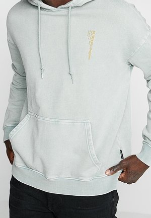 Hellgrauer Kapuzenpullover mit Fronttasche und kleinem gelbem Textlogo auf der Brust, getragen von einer Person mit den Händen nahe der Tasche.