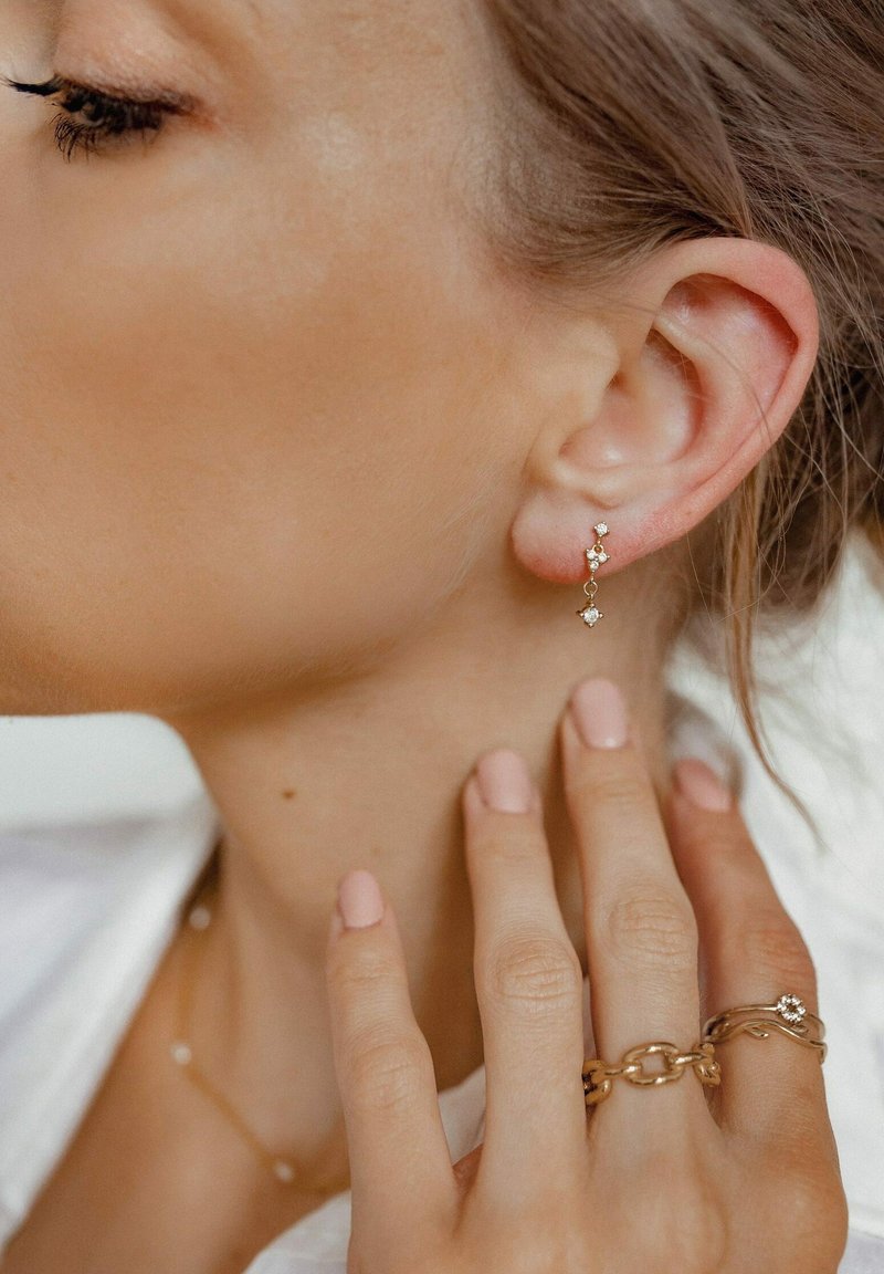 Pendientes de oro con tres elementos circulares y pequeños acentos de piedras, colgados de la lóbulo; la modelo se toca el cuello con uñas manicuras en tono nude-rosa.