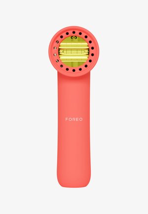 Foreo-Gesichtsgerät in Koralle mit rundem Kopf und gelber Innenfläche, ausgestattet mit Belüftungslöchern und einem glatten, ergonomischen Griff.