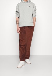 Persona que lleva una sudadera ligera gris de Nike, pantalones de pana marrones, zapatillas blancas, con tatuajes visibles en el antebrazo izquierdo, contra un fondo sencillo.