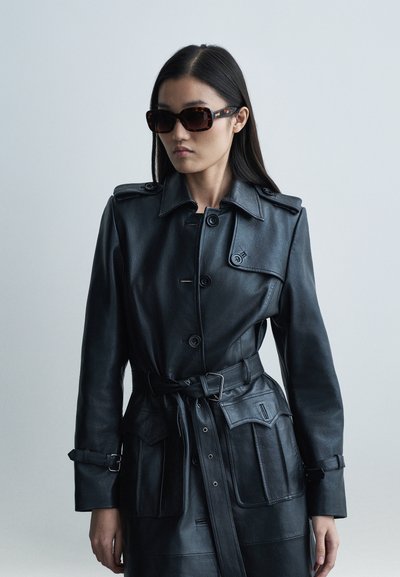 Juoda odinė tranšėjos striukė su diržu liemens srityje, petiniais epauletais ir didelėmis priekinėmis kišenėmis; derinama su tamsiais vėžlio kiauto spalvos akiniais nuo saulės.