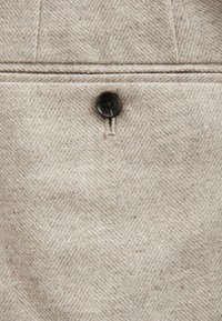 Beige herringbone-kangas, jossa on tekstuuri, ja tumma muovinen nappi, jossa on neljä reikää, säädetty beigeillä langoilla.
