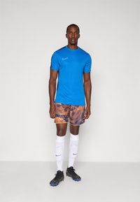 Blå atletisk T-skjorte med svarte detaljer, mønstrede oransje shorts, hvite sokker og svarte fotballsko, vist mot en ensfarget bakgrunn.