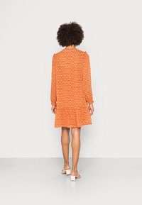 Vestido laranja com bolinhas brancas, mangas compridas, bainha com franzido e fecho de botão nas costas. O tecido é leve e fluido.