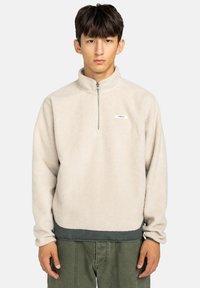 Polarowy pulower w jasnym beżu z półzamkiem, wysokim kołnierzem i kontrastowym, ciemnozielonym wykończeniem. Posiada małe logo na klatce piersiowej.